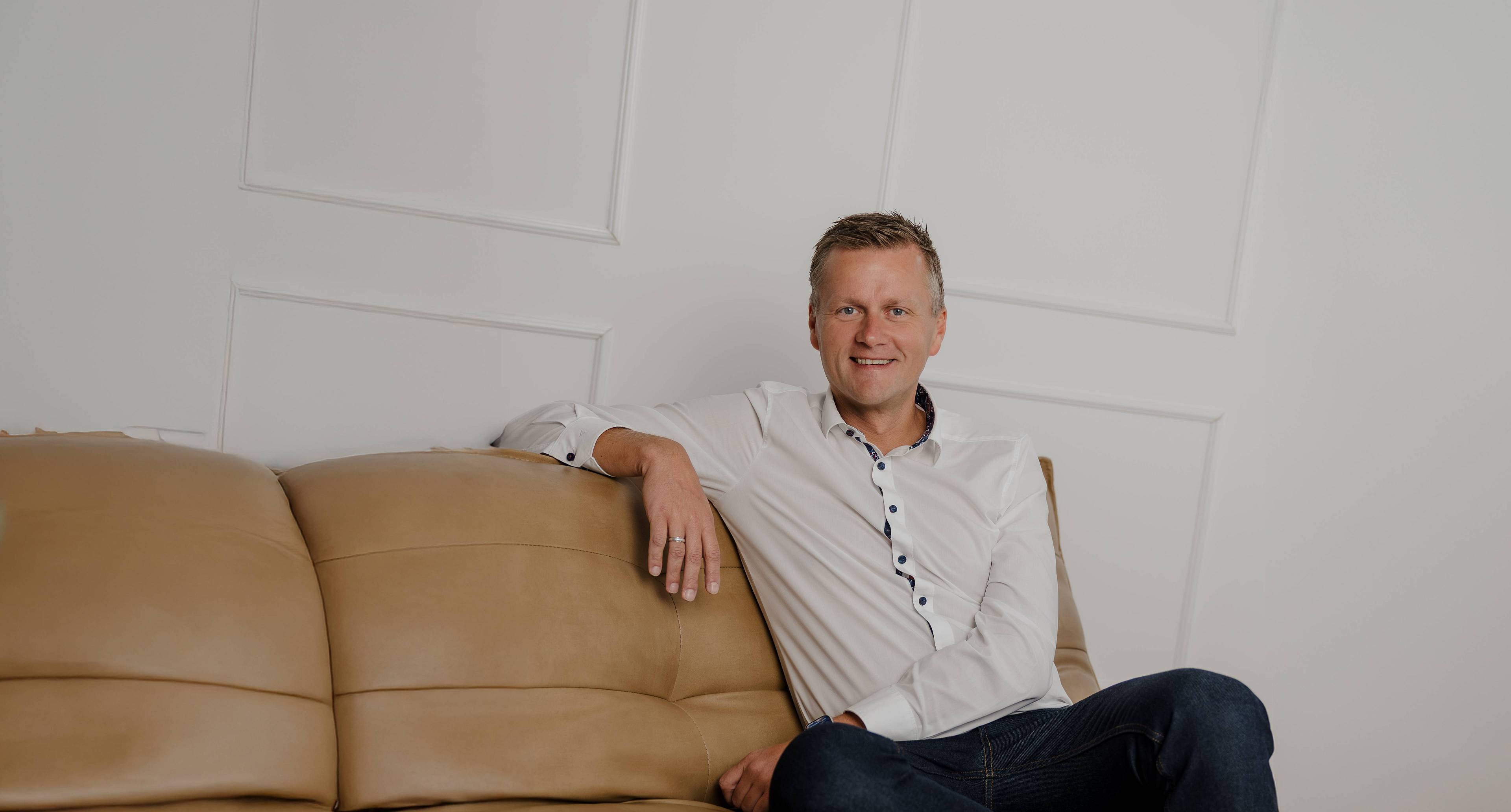 Das Bild zeigt Peter Vogt entspannt auf einem beigen Ledersofa. Mit offenem Lächeln, weißem Hemd und moderner Uhr vermittelt er Vertrauen, Kompetenz und Bodenständigkeit – ein authentisches Porträt, das seine Professionalität und Nahbarkeit unterstreicht.