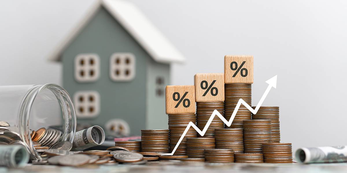 Aufgestapelte Münzen mit Prozentzeichen und steigender Kurve vor einem Modellhaus – Symbolbild für Immobilienfinanzierung und Zinsentwicklung.