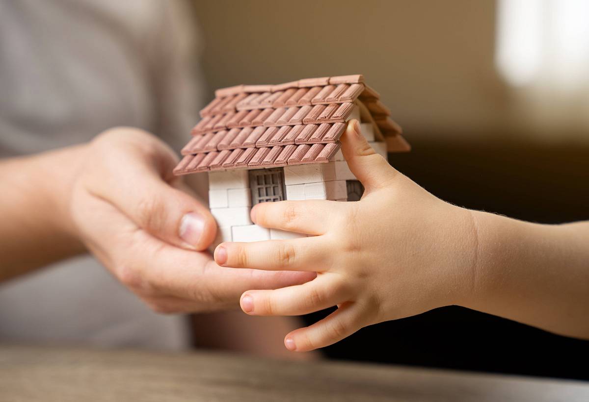 Erwachsene Hand übergibt einem Kind ein Miniaturhaus – symbolisch für Immobilienerbe und Generationenwechsel.
