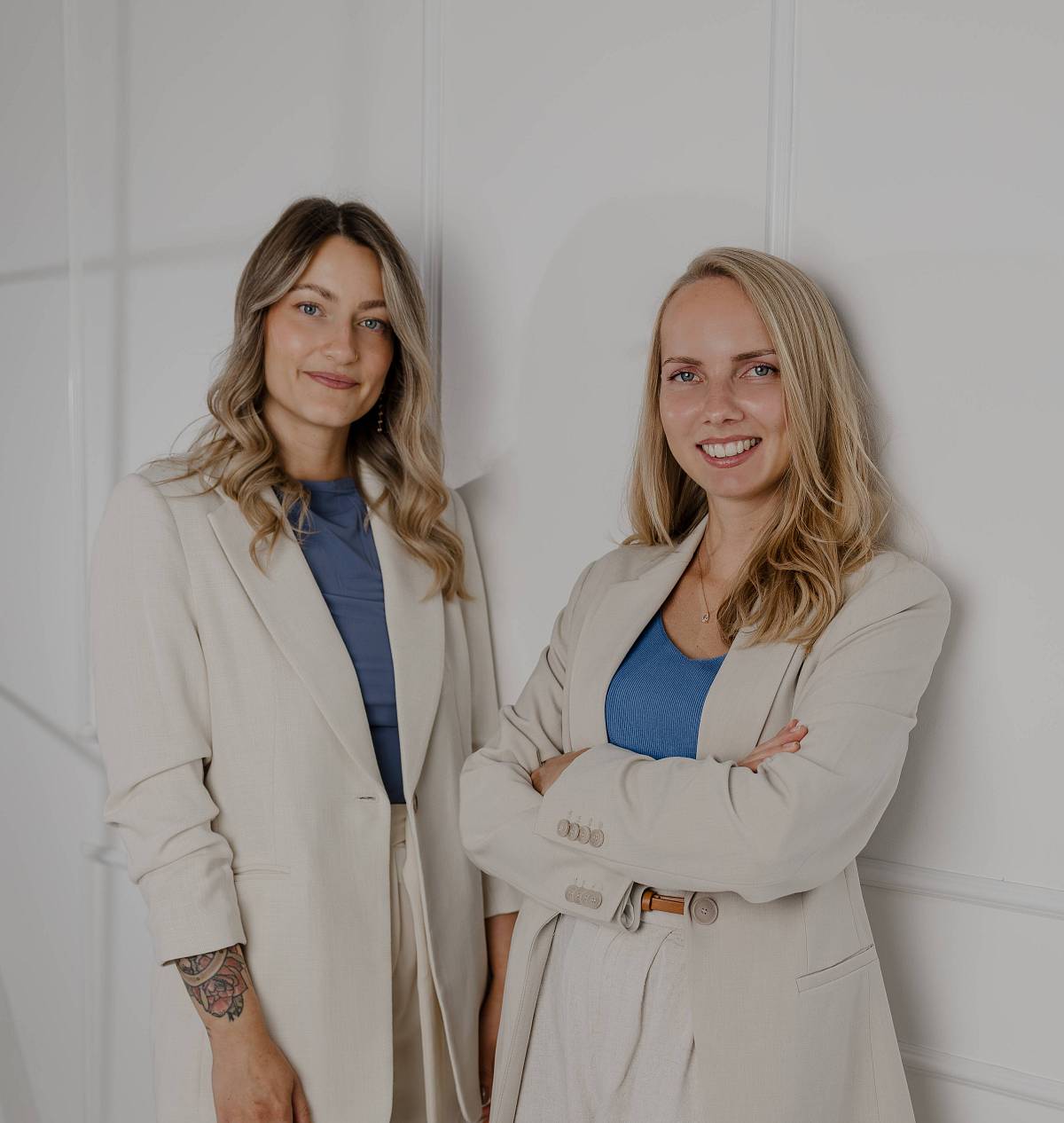 Das Bild zeigt zwei Mitarbeiterinnen von Peter Vogt Immobilien, die selbstbewusst und sympathisch vor einer hellen Wand stehen. In hellen Anzügen und mit blauen Akzenten verkörpern sie Professionalität, Kompetenz und den modernen Stil des Unternehmens.