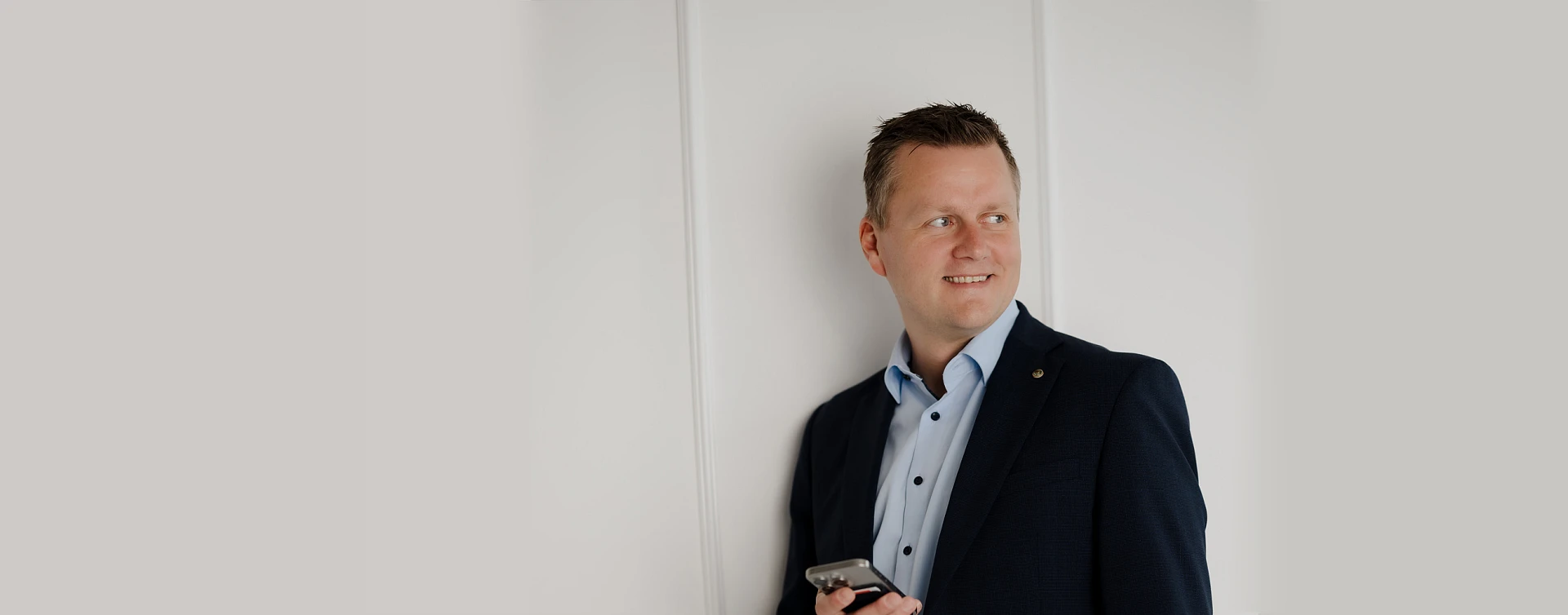 Mann in Business-Outfit lehnt entspannt an einer weißen Wand und blickt zur Seite, während er ein Smartphone in der Hand hält.