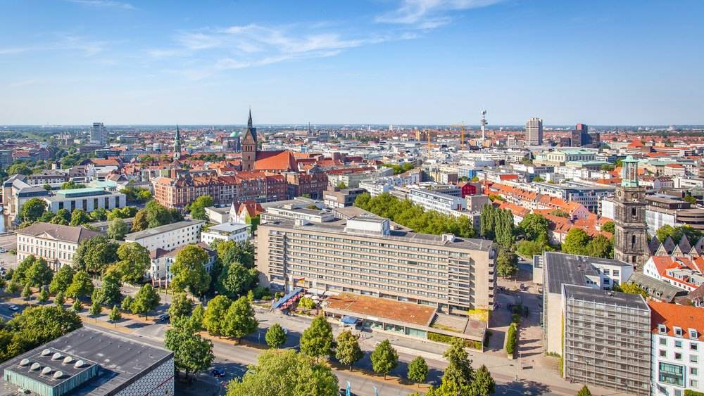 Panorama-Blick über die Innenstadt von Hannover mit markanten Gebäuden, grünen Baumreihen und blauem Himmel.