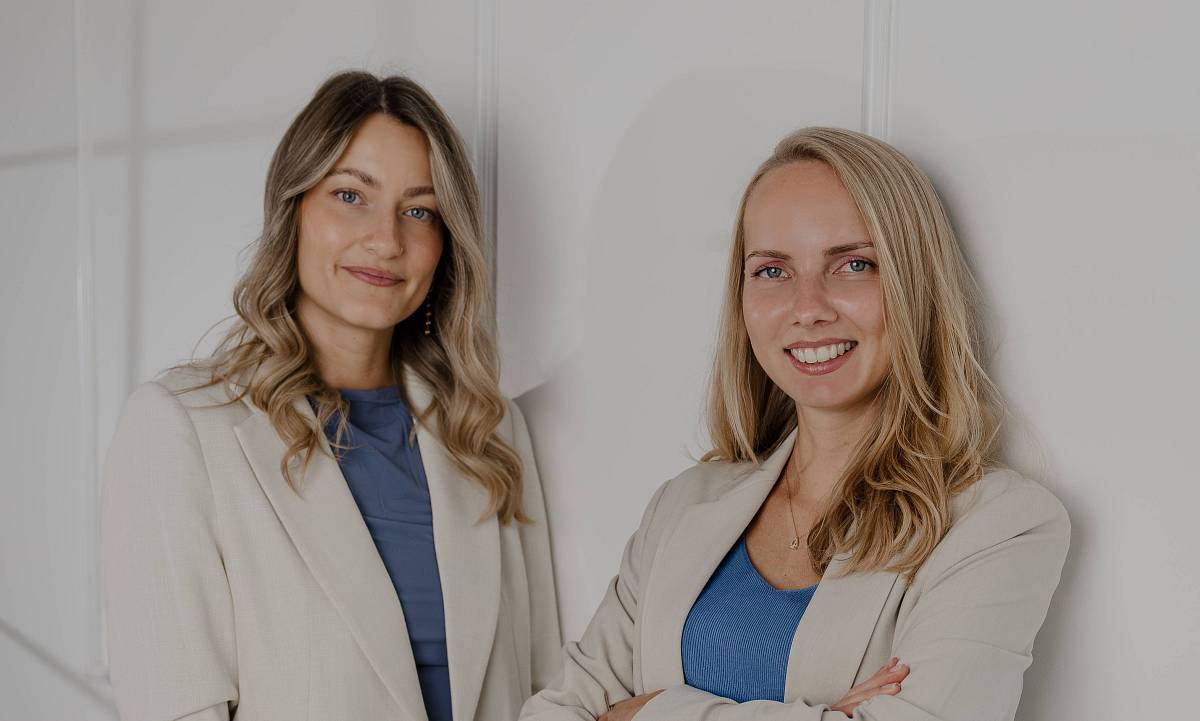 Das Bild zeigt zwei Mitarbeiterinnen von Peter Vogt Immobilien, die selbstbewusst und sympathisch vor einer hellen Wand stehen. In hellen Anzügen und mit blauen Akzenten verkörpern sie Professionalität, Kompetenz und den modernen Stil des Unternehmens.