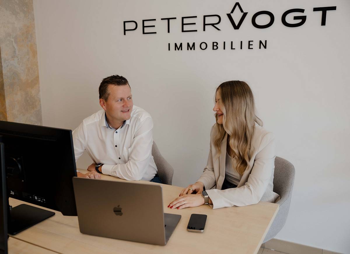 Das Bild zeigt Peter Vogt und seine Mitarbeiterin, die selbstbewusst und sympathisch im Büro sitzen. In hellen Anzügen und mit beigen Akzenten verkörpern sie Professionalität, Kompetenz und den modernen Stil des Unternehmens.