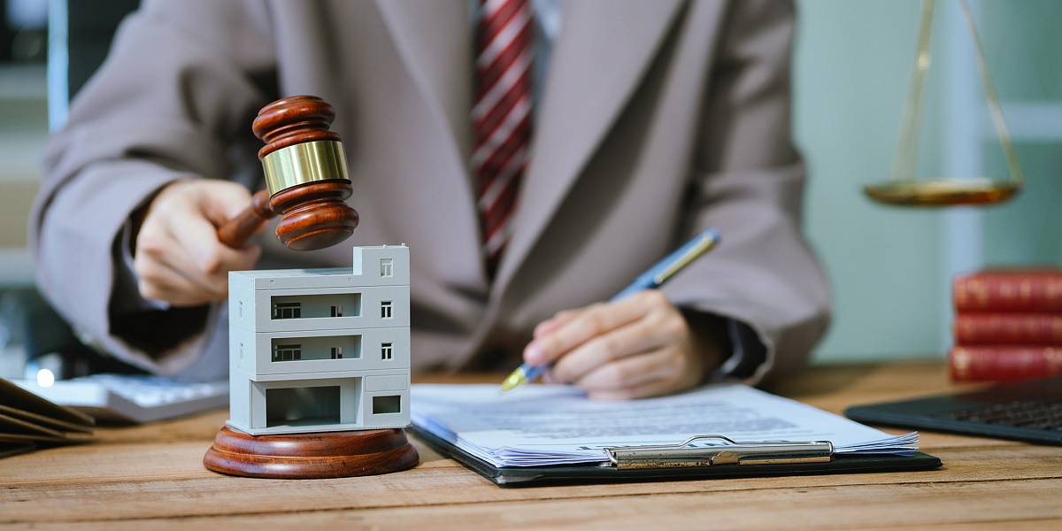 Berater sitzt am Schreibtisch und entscheidet mit Richterhammer neben Modell eines Mehrfamilienhauses über eine Immobilienverkaufsstrategie.