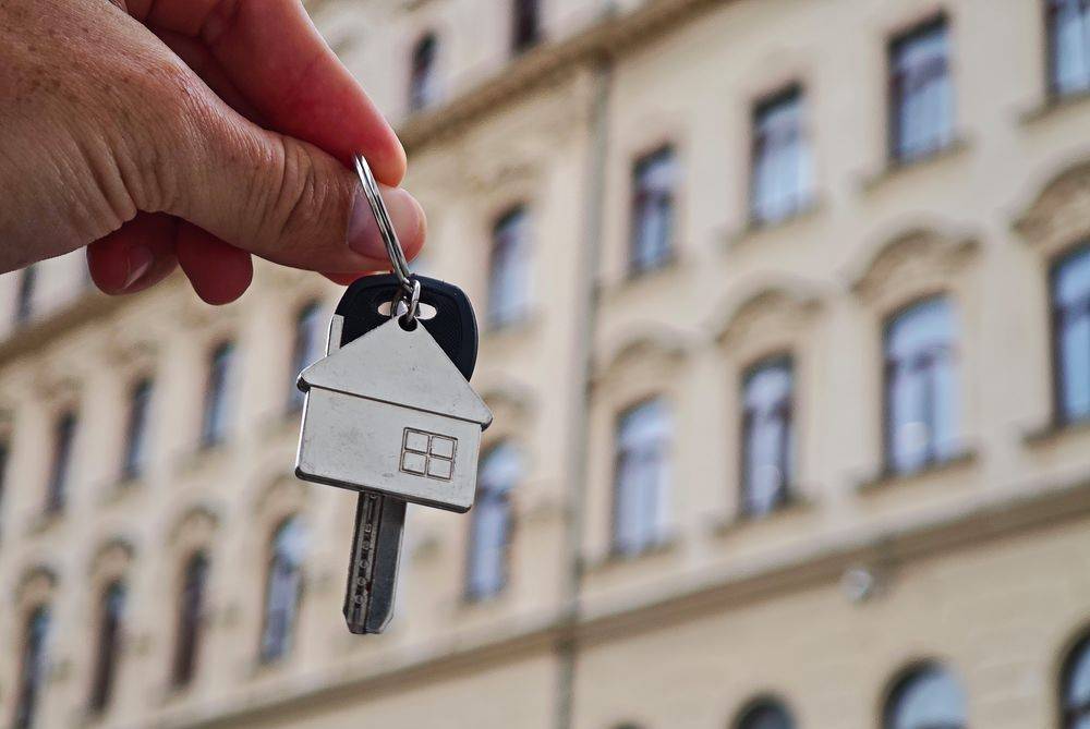 Schlüssel mit Hausanhänger vor der Fassade eines Mehrfamilienhauses als Symbol für Immobilienkauf oder -verkauf.