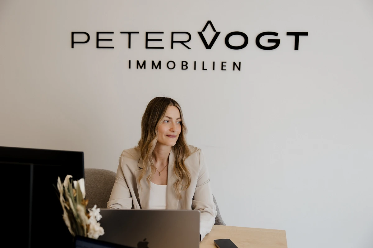 Mitarbeiterin von Peter Vogt Immobilien sitzt an einem Schreibtisch mit Laptop im Büro unter dem Firmenlogo.