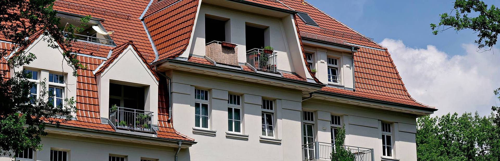 Mehrstöckiges Wohnhaus mit rotem Ziegeldach, Gauben und Balkonen unter blauem Himmel.