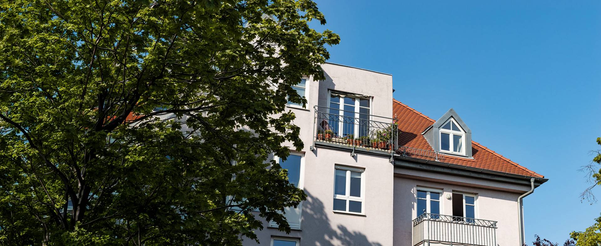 Mehrstöckiges Wohnhaus mit Balkonen, rotem Ziegeldach und großen Bäumen vor blauem Himmel.