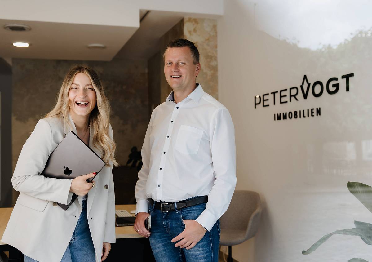 Peter Vogt und eine Mitarbeiterin stehen lächelnd im Büro von Peter Vogt Immobilien.