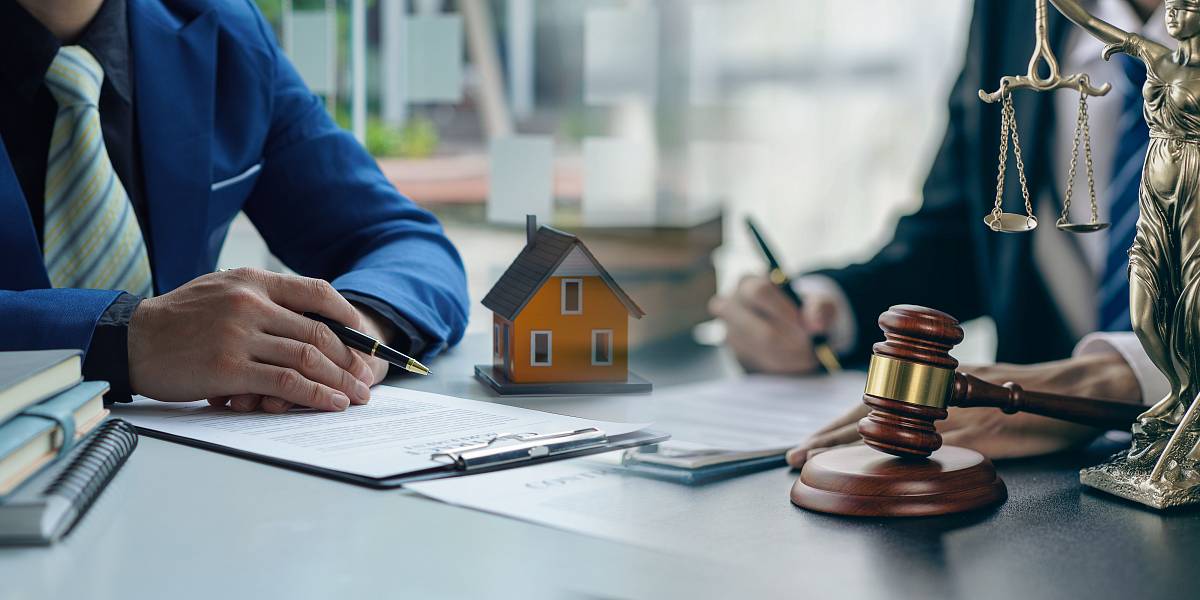 Zwei Personen am Schreibtisch mit Gesetzbuch, Justitia-Figur und Modellhaus – rechtliche Aspekte bei Immobilientransaktionen.