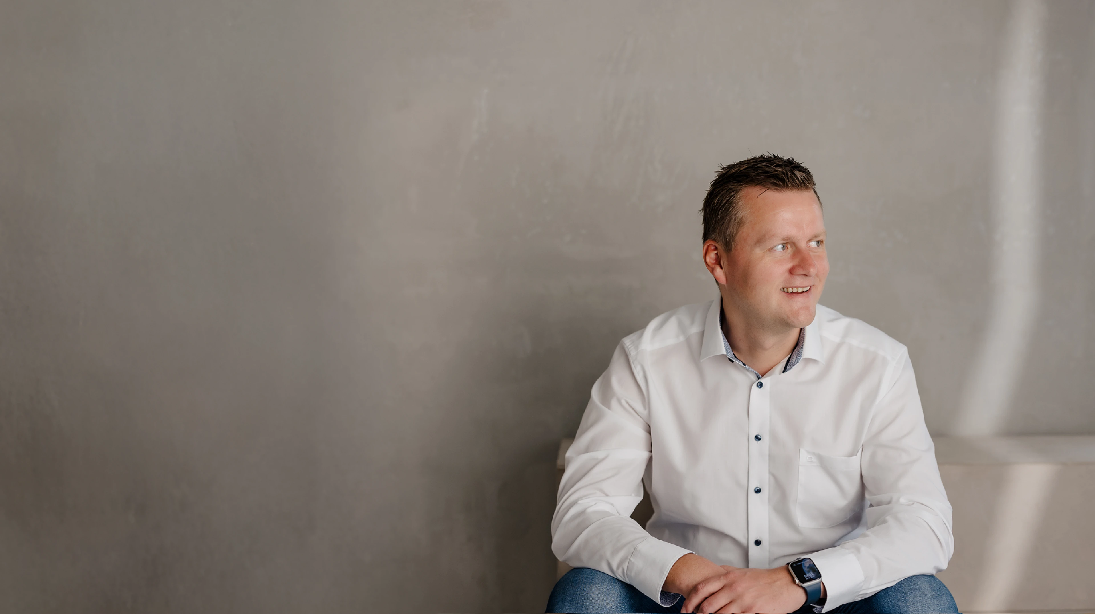 Immobilienexperte in weißem Hemd sitzt vor grauer Wand und blickt zur Seite