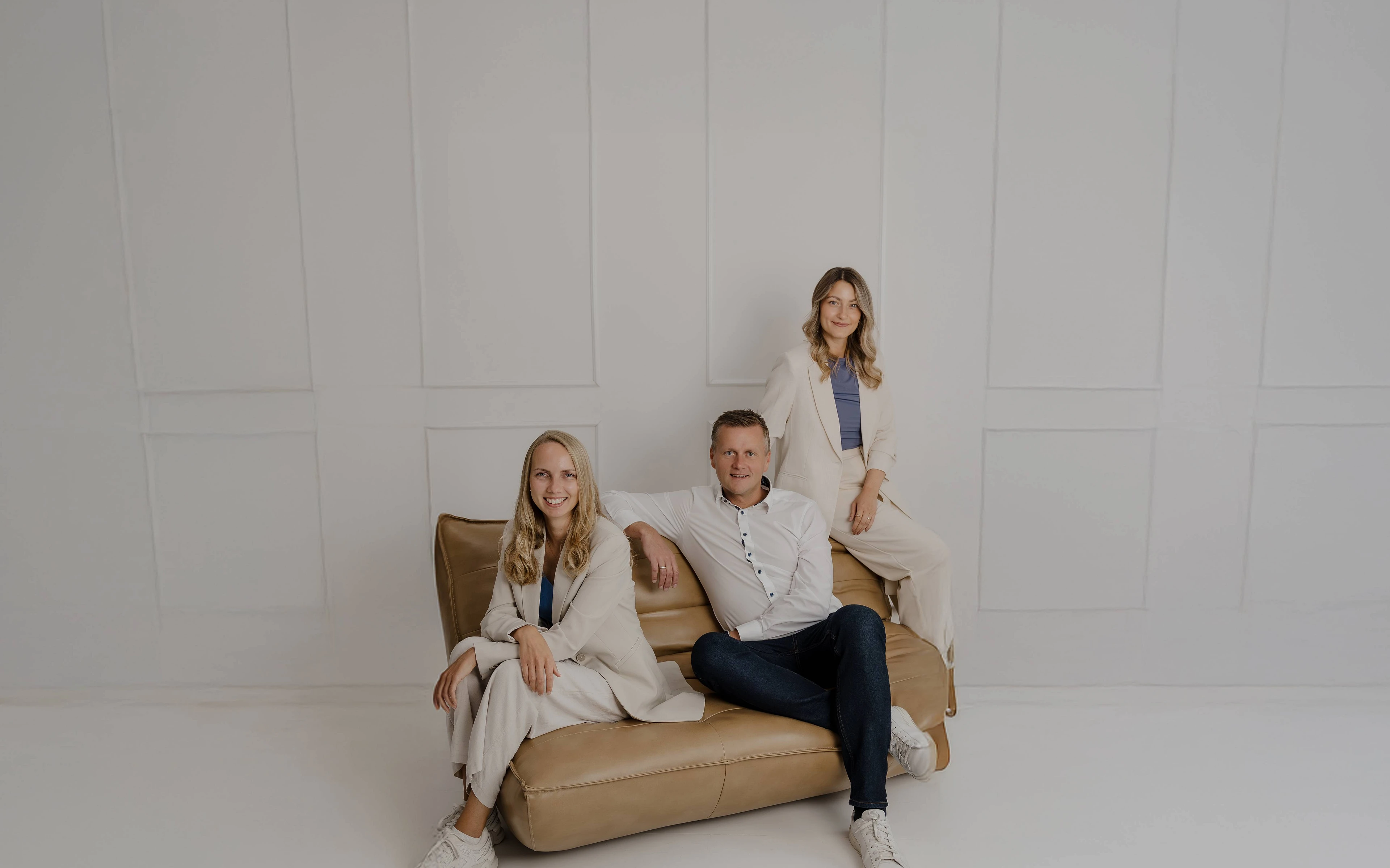 Das Bild zeigt Peter Vogt mit zwei Mitarbeiterinnen in einem hellen, modernen Studio. Sie sitzen und stehen locker auf einem beigen Sofa vor einer weißen Wand – ein professionelles, aber nahbares Porträt, das Seriosität und Sympathie ausstrahlt.