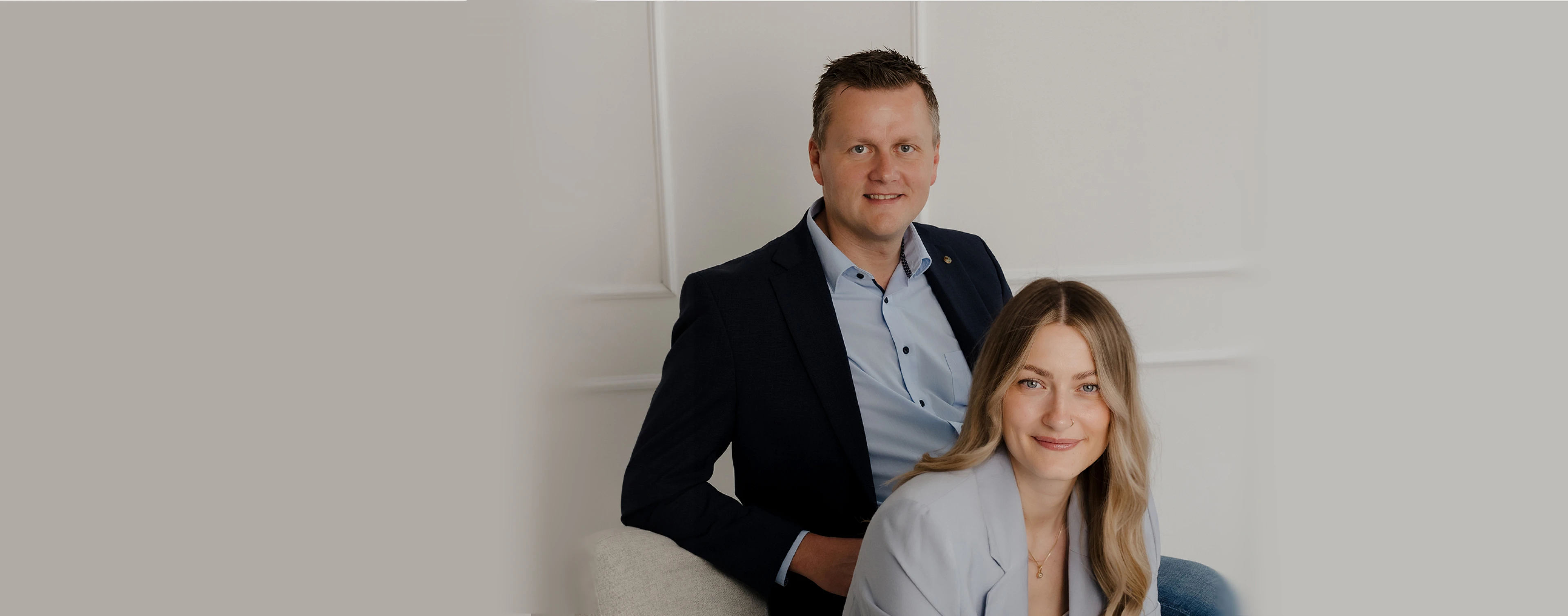 Peter Vogt und Leijla von Peter Vogt Immobilien sitzen gemeinsam vor heller Wandkulisse und blicken freundlich in die Kamera.