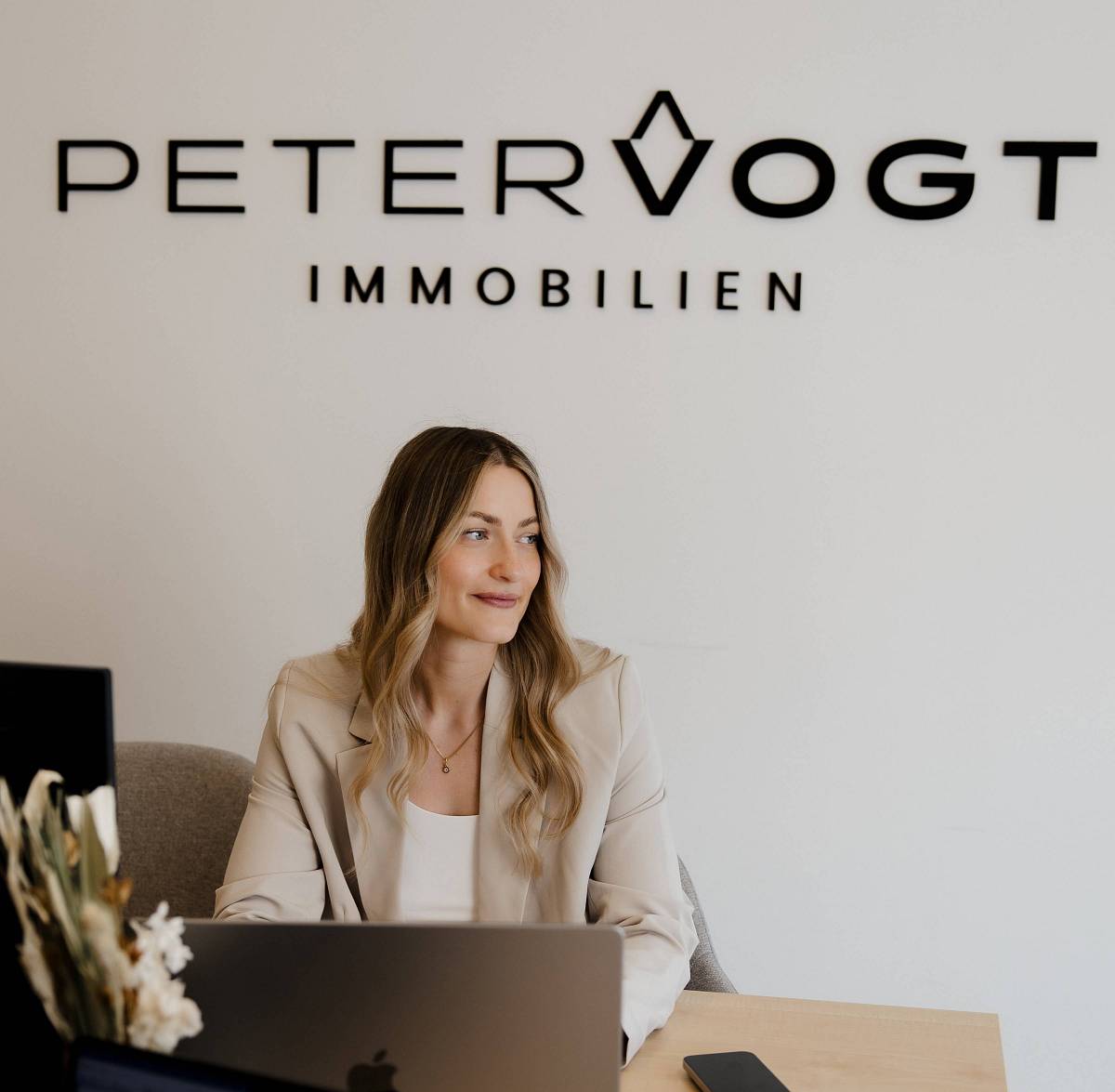 Junge Frau im Büro von PETER VOGT IMMOBILIEN sitzt an einem Schreibtisch mit Laptop und Smartphone, freundlich lächelnd – symbolisiert kompetente Immobilienberatung.