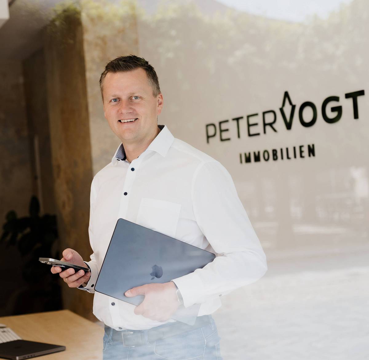 Peter Vogt steht mit Laptop und Smartphone in der Hand vor dem Schaufenster seines Immobilienbüros – freundlich und professionell.